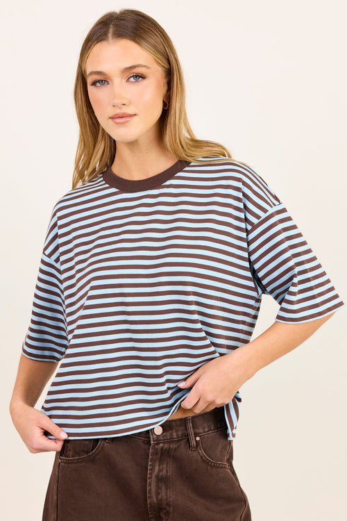 A woman modeling a brown and white striped short-sleeve t-shirt with a solid brown crew neck, paired with dark brown jeans, against a plain off-white background.