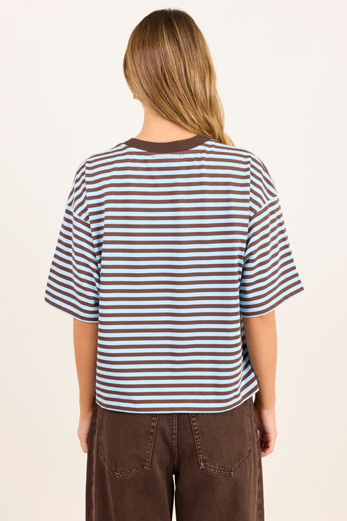 A woman viewed from behind wearing a brown and white horizontally striped short-sleeve t-shirt and brown pants, set against a plain off-white background.