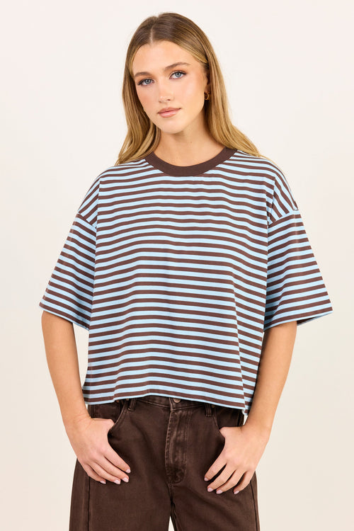 A young woman modeling a short-sleeve crewneck t-shirt with horizontal brown and white stripes, paired with dark brown pants, against a plain off-white studio background.