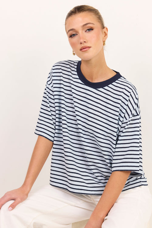 A woman wearing an oversized short-sleeve striped t-shirt with navy blue horizontal stripes on a white background, paired with white pants, against a plain off-white studio background.