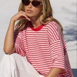 A woman wearing a red and pink striped t-shirt and white pants sits on the edge of a swimming pool, wearing sunglasses.