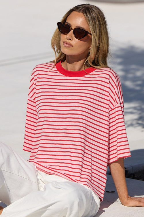 A woman wearing a red and white striped t-shirt and white pants sits on a white tiled poolside deck next to a tree.