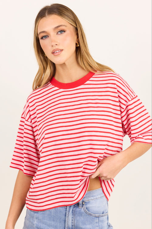 A woman modeling a red and white horizontally striped short-sleeve crop top with a solid red crew neck, paired with light blue jeans, against a plain off-white background.