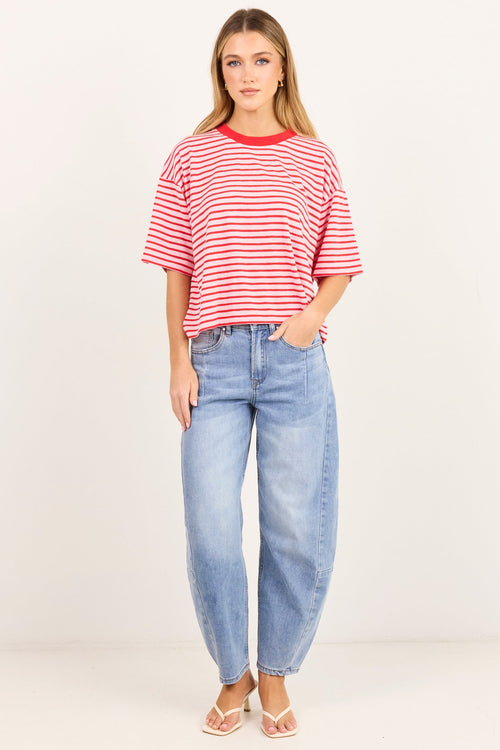 A woman modeling a red and white horizontally striped short-sleeve crop top with red trim, paired with light-wash blue jeans and white sandals, standing against a plain off-white studio background.