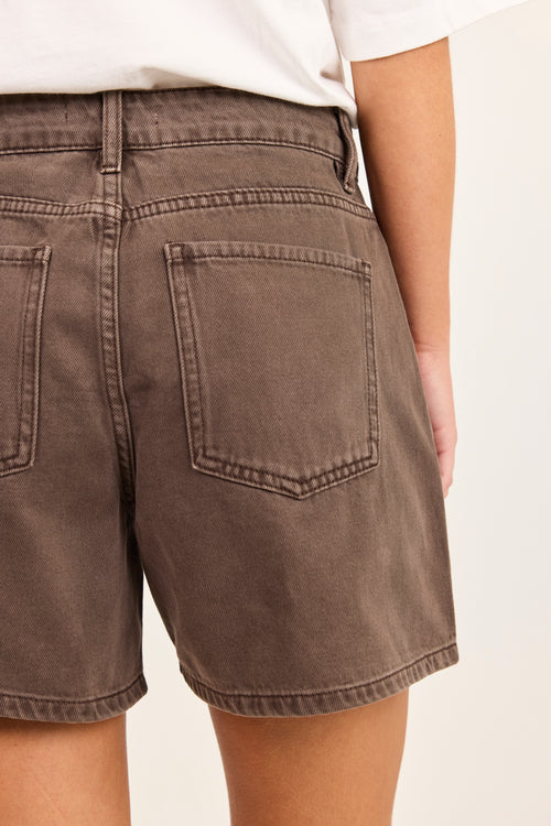 Close-up back view of a person wearing brown denim shorts with a white top, showing the back pockets and waistband details against a plain light background.