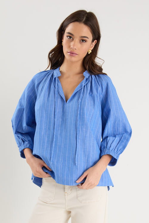 Woman wearing a relaxed-fit, blue pinstripe top with mid sleeves and a split neckline, styled with off-white jeans.