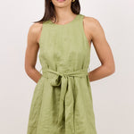 A woman modeling a sleeveless olive green linen dress with a matching tie belt, set against a plain light beige studio background.