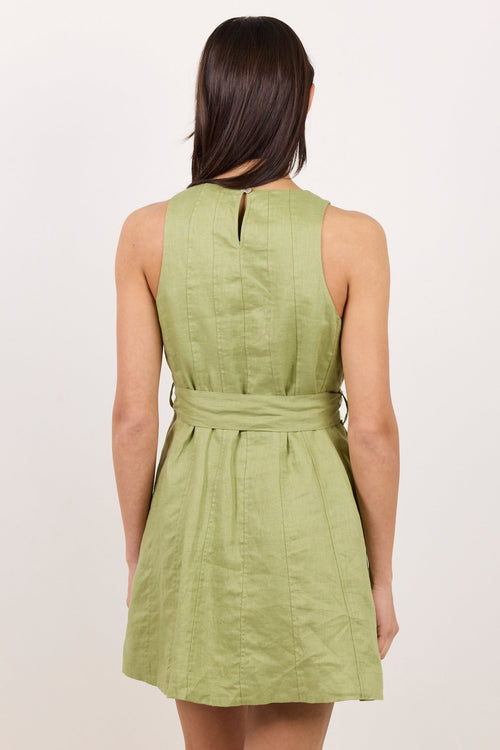 A woman viewed from the back wearing a sleeveless light green dress with a tie waist, against a plain off-white studio background.