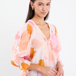 Woman wearing a pink and orange abstract printed short sleeve top with a V-neckline, tie front, and relaxed fit, paired with light blue jeans.