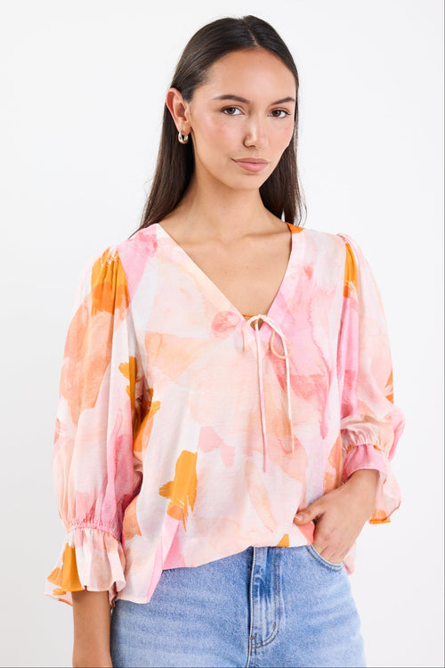 Woman wearing a pink and orange abstract printed short sleeve top with a V-neckline, tie front, and relaxed fit, paired with light blue jeans.