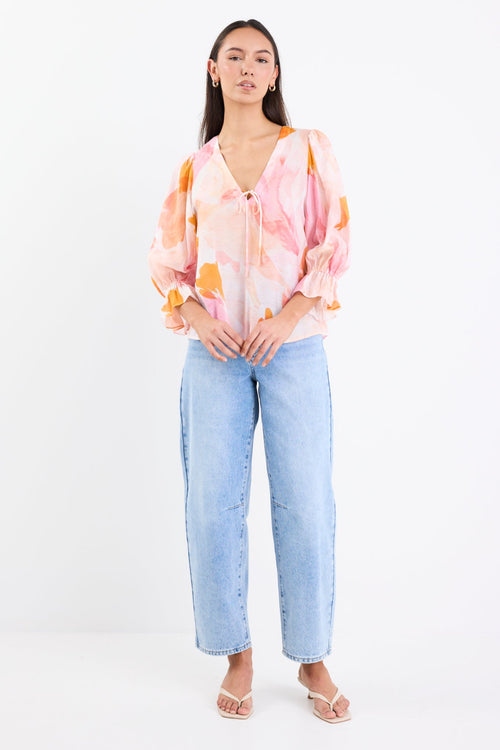Woman wearing a pink and orange abstract printed short sleeve top with a V-neckline, tie front, and relaxed fit, paired with light blue jeans.