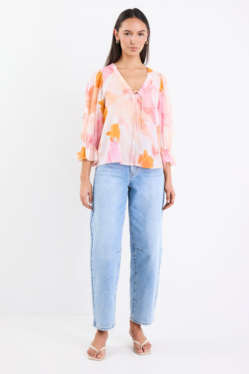 Woman wearing a pink and orange abstract printed short sleeve top with a V-neckline, tie front, and relaxed fit, paired with light blue jeans.