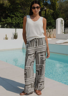 Woman standing by a pool wearing a white top and patterned pants.
