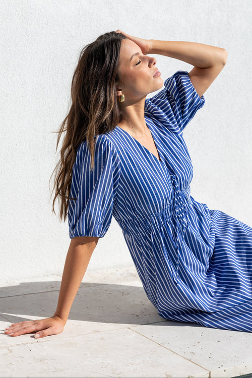 A woman wearing a blue and white vertically striped midi dress with puff sleeves and a gathered waist, posing on a light stone surface against a white textured wall.