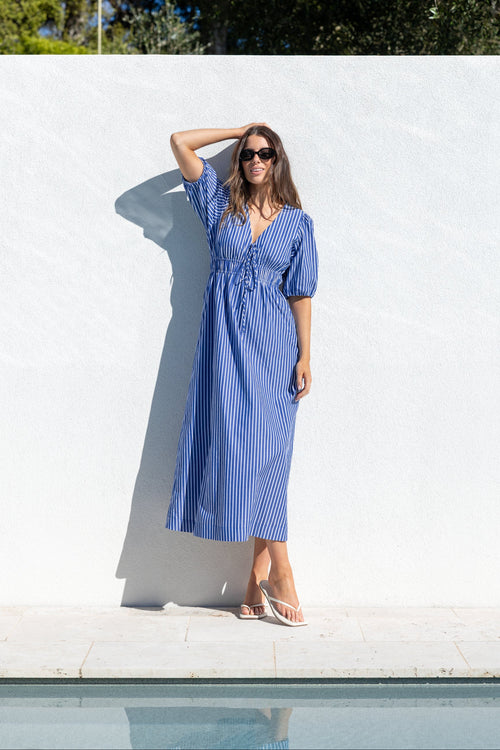 A woman wearing a blue and white vertically striped midi dress with puff sleeves and a tie waist, posing outdoors against a white wall near a pool.