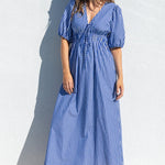 A woman wearing a blue and white vertically striped midi dress with puff sleeves and a V-neckline, standing against a white wall on a sunny day.