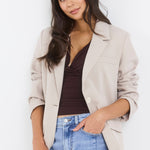 Woman wearing a beige blazer, brown top, and blue jeans on a white background