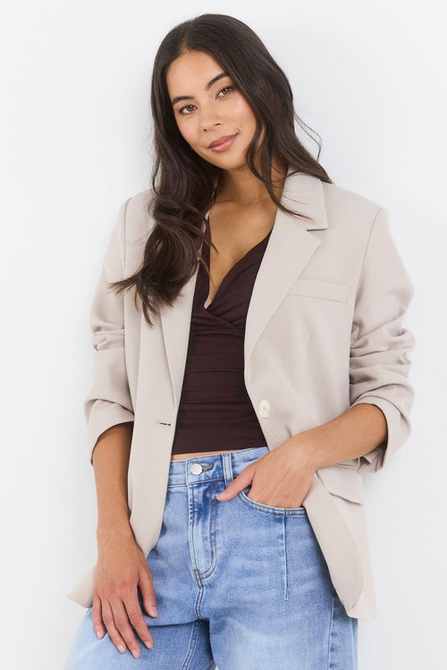 Woman wearing a beige blazer, brown top, and blue jeans on a white background