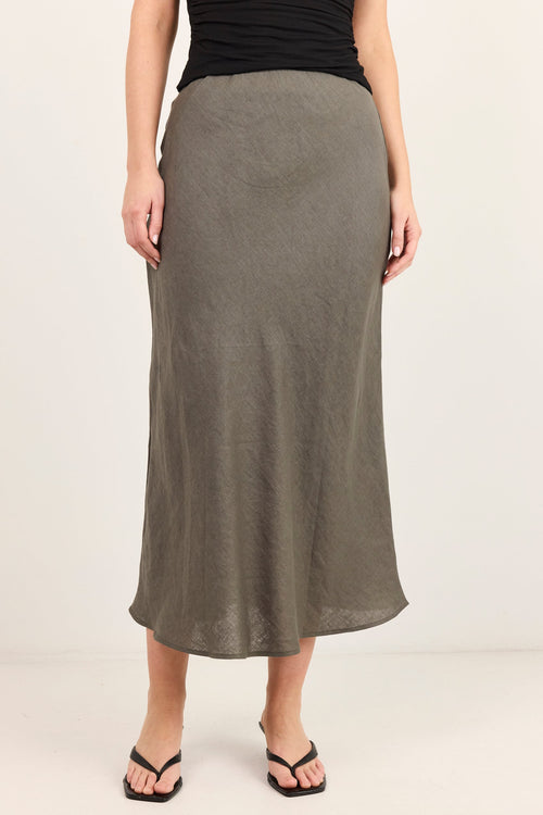 A woman modeling a long, flowing midi skirt in a muted olive-gray color, paired with a black sleeveless top and black thong sandals, against a plain off-white studio background.