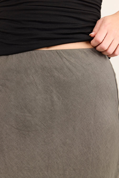 Close-up of a person wearing a dark gray skirt or wrap skirt, pulling the fabric outward with one hand to show its drape and texture. A black top is also visible.