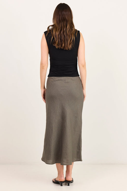 A woman viewed from behind wearing a sleeveless black top and a long, straight, ankle-length olive green skirt, standing against a plain off-white studio background.