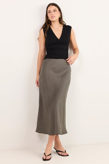 A woman modeling a sleeveless black wrap-style top and a long, straight, knee-length olive green skirt, paired with black flip-flop sandals, against a plain off-white studio background.