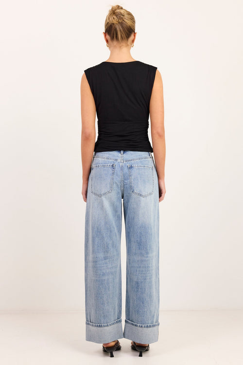 A woman viewed from behind wearing a black sleeveless top and light-wash wide-leg denim jeans with rolled cuffs, standing against a plain light background.