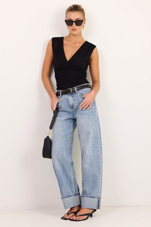 A woman modeling a black sleeveless V-neck top, light-wash wide-leg jeans, black sandals, and sunglasses, holding a small black handbag against a plain white studio background.