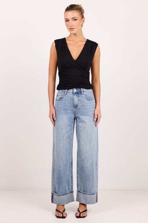 A woman modeling a black sleeveless wrap-style top and light-wash wide-leg jeans with cuffed hems, standing against a plain white studio background.