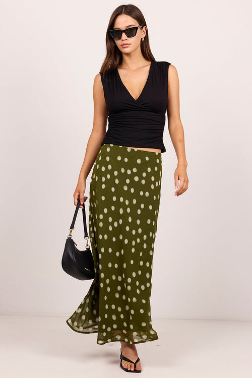 A woman modeling a sleeveless black wrap-style top and a flowing olive green skirt with white polka dots, holding a black handbag against a plain light gray studio background.