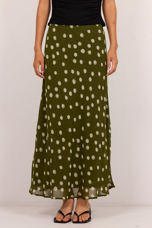 An olive green maxi skirt with a white polka dot pattern, worn by a model with a black top and black sandals against a plain white studio background.
