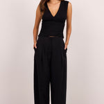 A woman modeling a black sleeveless wrap-style top and matching black wide-leg trousers, standing against a plain light studio background.