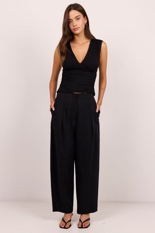 A woman modeling a black sleeveless wrap-style top and matching black wide-leg trousers, standing against a plain light studio background.