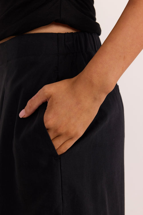 Close-up of a person wearing black wide-leg trousers with a side pocket, shown from the waist down with one hand in the pocket.