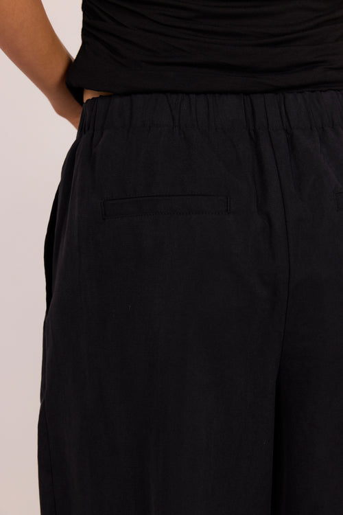Close-up of a person wearing black wide-leg pants with an elastic waistband and a side welt pocket, paired with a black top against a plain light background.