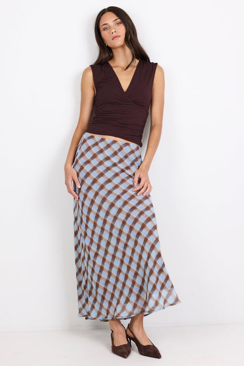 A woman modeling a sleeveless dark brown wrap-style top and a long brown, white, and light blue gingham skirt, paired with dark brown heels, against a plain white studio background.
