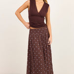 A woman modeling a sleeveless dark brown wrap-style top and a matching dark brown maxi skirt with small white polka dots, standing against a plain off-white studio background.