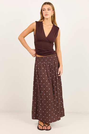 A woman modeling a sleeveless dark brown wrap-style top and a matching dark brown maxi skirt with small white polka dots, standing against a plain off-white studio background.