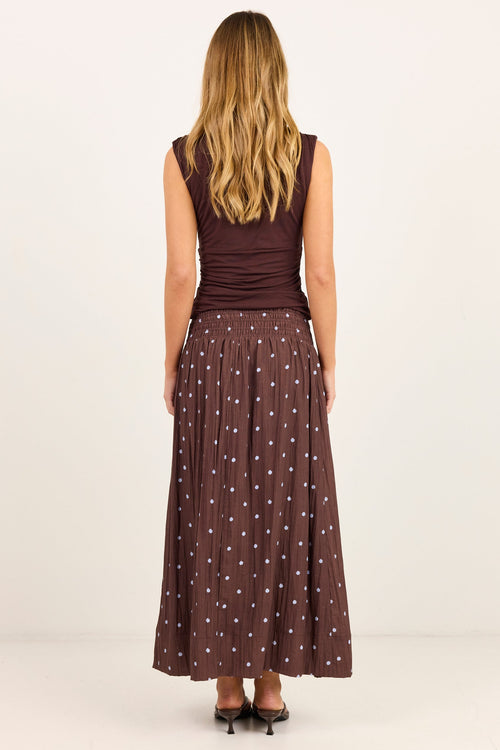 A woman viewed from the back wearing a sleeveless dark brown top and a matching long pleated skirt with small white polka dots, standing against a plain light studio background.