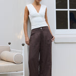 A woman stands outdoors against a white wall wearing a white sleeveless top and dark brown wide-leg trousers, with sandals on a patio next to a white chair.