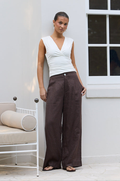 A woman stands outdoors against a white wall wearing a white sleeveless top and dark brown wide-leg trousers, with sandals on a patio next to a white chair.