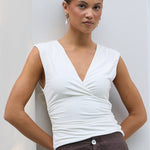 A woman stands indoors against a white wall next to a window, wearing a white sleeveless wrap-style top and dark brown wide-leg pants. A light-colored cushioned chair is visible on the left.