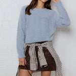 Woman wearing a light blue sweater and striped skirt with a brown handbag against a white wall.