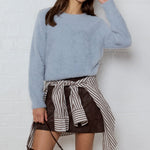 A woman modeling a light blue fuzzy long-sleeve sweater, a brown mini skirt with a black and white striped shirt tied around the waist, and a dark brown leather shoulder bag, standing against a white brick wall.
