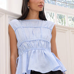 A woman wearing a light blue sleeveless peplum top with gathered detailing and black wide-leg pants, standing in a bright white room with decorative wall molding and a window.