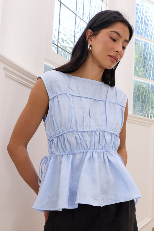 A woman wearing a light blue sleeveless smocked blouse with a peplum hem and black trousers, standing in a bright white interior with windows and wooden stairs.
