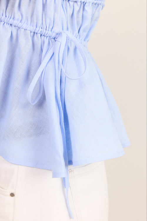 Close-up of a light blue peplum-style top with an elasticized gathered waist and a side tie, worn with white pants against a plain light beige background.
