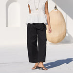 A woman stands by a swimming pool wearing a white sleeveless peplum top, black wide-leg cropped pants, and black sandals in a modern white outdoor setting with hanging woven chairs.