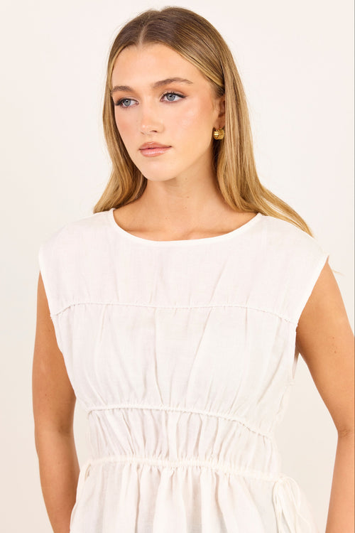 A woman with blonde hair wearing a white sleeveless dress with a gathered waist, photographed against a plain off-white background.