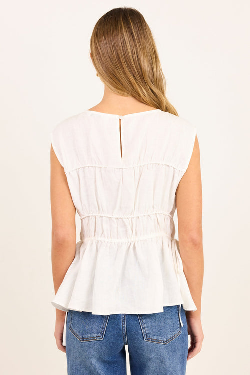 A woman viewed from the back wearing a white sleeveless peplum top with a smocked waist and a small back slit, paired with blue jeans against a plain off-white background.